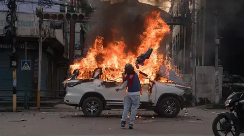 Getty Images A person draped in Nepal's national flag throws an obejct towards a flaming vehicle in the street
