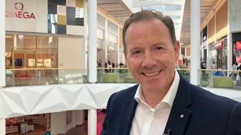 A smiling man with short, brown hair who is wearing a white shirt and blue suit jacket. In the background are several shops and shoppers.