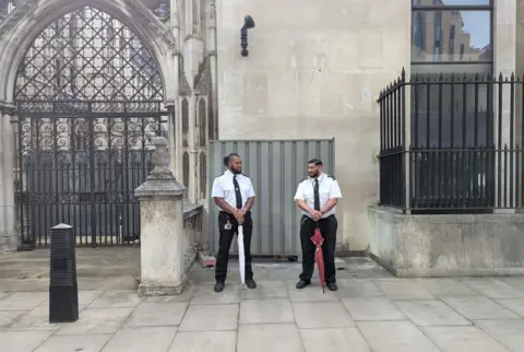 PA Media Two men holding furled golf umbrellas in front of a metal barrier in front of a wall. They are dressed in smart security guard outfits and look toward each other.