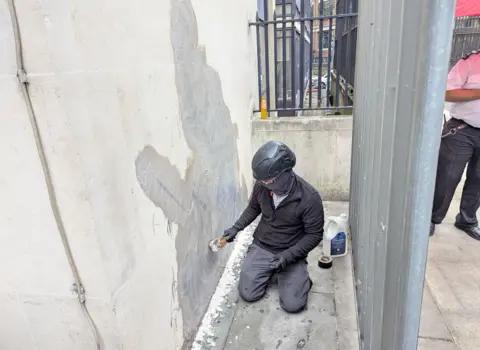 PA Media Man with covered face and a bike helmet removing the mural with white spirits