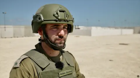BBC/Dave Bull Nadav Shoshani is wearing a khaki military uniform, including a helmet. His name is written on the protective vest across his chest. He appears to be in his 20s or 30s and has dark eyebrows and a beard.