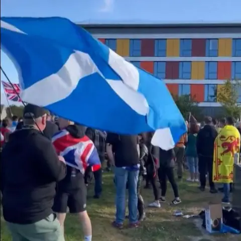 A protest outside the Hampton by Hilton hotel in Westhill, Aberdeenshire. A man carries a large saltire in the foreground. Another man is draped in a union jack, and a third has the lion rampant around his shoulders. All have their backs to the camera. 