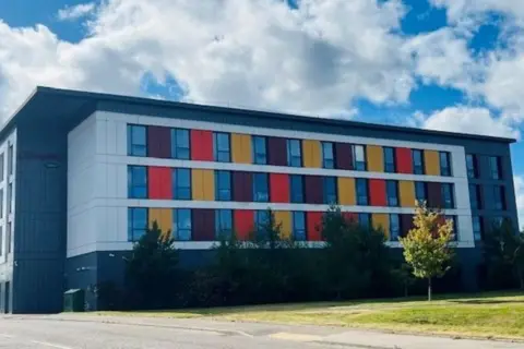 Hotel building with bright panels next to windows, and trees and grass outside, under a cloudy blue sky.