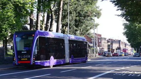 Pacemaker A purple single decker Glider bus on a road lined with trees, ahead on the road are a number of red brick houses and cars stuck at traffic lights. 