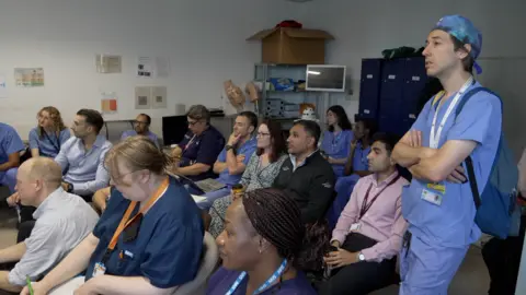 Medics gather in a busy meeting room - some are sitting - some are standing. There are about 16 people in the photo. Some are wearing surgical clothes, some in plain clothes