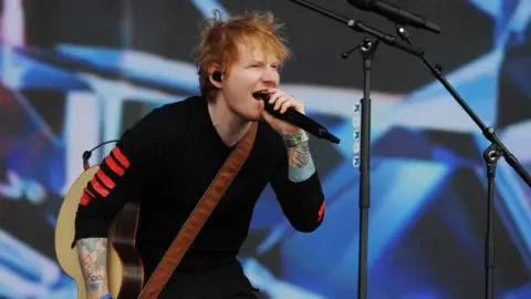 Sarah Jeynes/BBC Ed Sheeran on stage singing into a microphone and holding a brown semi-acoustic guitar.