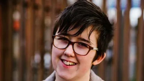 PA Media Lyra Mckee is smiling and wearing glasses in a grey coat