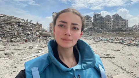 Plestia Alaqad A woman with brown hair looks at the camera. She is wearing a thick blue press jacket. Behind her is a large mound of rubble, as well as severely damaged buildings.