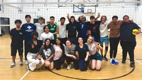 Konstantin Schmidt A group of students are standing in front of a volleyball net in a sports hall. They are all facing the camera.