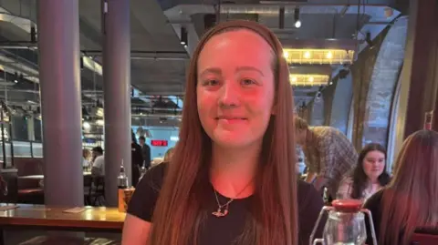 Handout Rebecca is sitting in a restaurant. She is smiling at the camera wearing a black T-shirt