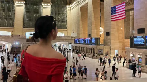 Tian Liu Tian is in a train station. She is looking away from the camera looking at an american flag on the wall. She is wearing a red top.