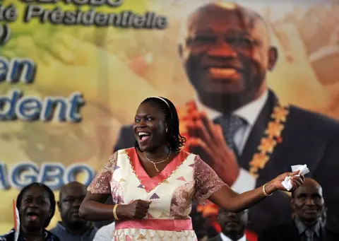 AFP via Getty Images Simone Gbagbo stands in front of a picture of her husband, wearing a pink and wife dress.