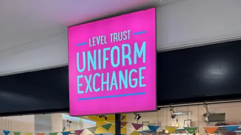 Kerri Porthouse A pink sign that reads "Level Trust Uniform Exchange" which is hanging above a glass-panelled shop front. Bunting can be seen inside the shop, hung from the roof.