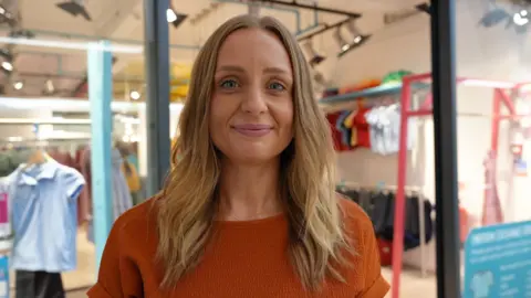 Dawid Wojtowicz/BBC Kerri Porthouse smiles at the camera as she stands outside a school uniform shop. She is wearing an orange top and has long brown hair.