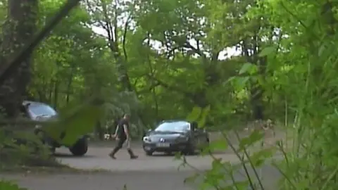 Gwent Police Still image of surveillance footage with a some foliage in the foreground and showing a man wearing black walking to a black car with a supermarket carrier bag in his hand. He is walking on tarmac and behind him and car is a dense woodland