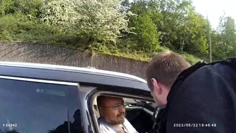 Gwent Police Still image of police bodycam footage of a man getting arrested in a dark coloured taxi by a police officer wearing black on a busy road.