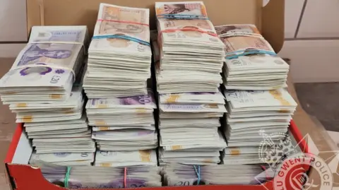 Gwent Police A picture of piles of banknotes bundled and secured with elastic bands in a red Nike shoe box. The lid can't be closed because the piles are so tall.