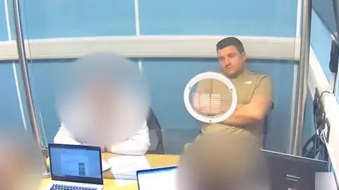 Gwent Police A man in police custody wearing a brown top is being questioned by two detectives who are sat in front of him, both using laptop computers. Alongside the arrested man is a man wearing a white shirt and his face is blurred out 