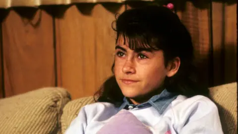 Getty Images Roxana Zal, pictured in 1984's Something About Amelia, sitting on a sofa, wearing a white jumper with her black hair tied up