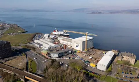 An aerial view of a shipyard. There are grey buildings, a yellow crane and a large ship berthed on the quayside