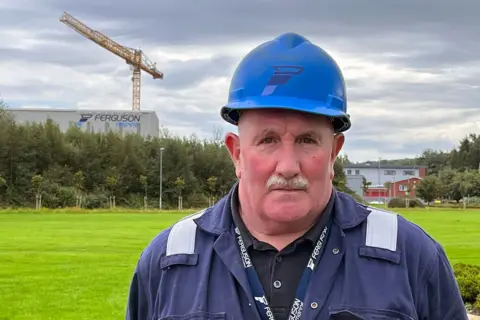 A man with a grey moustache and a blue helmet, wearing a blue boilersuit. In the background is a large yellow crane and a large grey building with the workds Ferguson Marine on the side. 