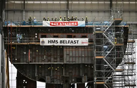 Getty Images A large warship, under construction, with workers on the deck and scaffolding at the side. The ship is in profile so the interior compartments are visible