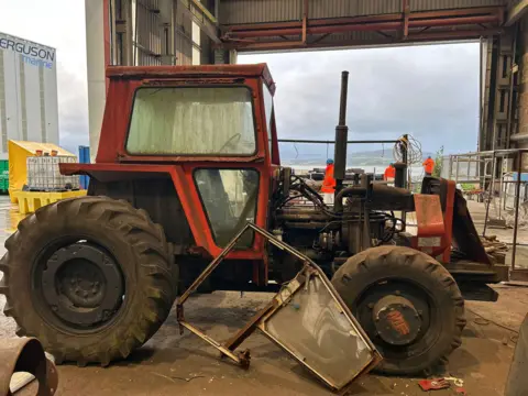 
A red tractor in a fabrication shed. One of the doors of the tractor has fallen off and is propped up against the vehicle