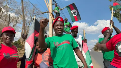 Supporters of Lazarus Chakwera's MCP on the roadside. The main figure in the centre is holding his fist up and wearing a green party T-shirt and red beret. Others are waving party flags.