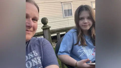 Family handout A selfie of Juile and Rosie. They are both sitting outside and there is a static caravan in the background. Julie is on the left of frame and only part of her face and body can be seen. She is smiling. Rosie is on the right of frame and is looking down the camera. She has brown, straight hair that is falling below her shoulders. Both Rosie and Julie are wearing T-shirts.