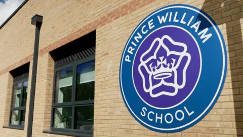 DJ McLaren/BBC An external brick wall with a large circular Prince William School logo. The middle of the logo is a purple circle, which has a white crown and white rose-style motif in the middle. The words "Prince William School" are written in white against a blue background, which runs around the central circle. Two windows and a drainpipe can be seen further along the wall in the background. 