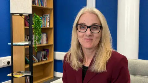 Ben Schofield/BBC Dr Joanne Summers looking direct to camera. She is sitting inside a room with deep blue walls and a white pillar over her shoulder to the right. On the left behind her is a bookshelf partially-filled with books, as well as a green plant. Dr Summers is smiling and is wearing a dark red jacket over a black top. She has black rimmed, rectangular glasses on and her blonde hair is parted in the middle and is falling over her shoulders.
