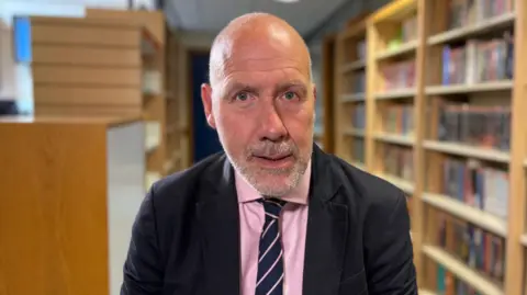 Ben Schofield/BBC Geoff Barton looking direct to camera. He is sitting down and rows of filled bookshelves are visible behind him, though are out of focus. He is wearing a dark jacket, pink shirt and blue and pink diagonally striped tie. Geoff is mostly bald but has a greying beard. He has blue eyes.