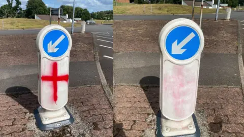 Sam Higgins Photograph shows a collage side-by-side of a white road bollard. The left photo shows the England flag upon the bollard, while the right hand side photo shows it after it has been scrubbed off.