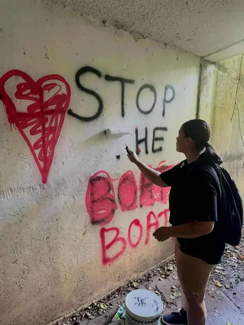 Contributor photo Photograph shows a young woman, wearing a black t-shirt and black shorts, holding a paint brush roller to a subway wall. On the wall, spray painted lettering reads: "Stop he Boats."