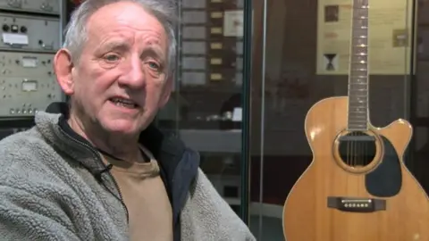A man with white hair, wearing a grey fleece and brown top. Next to him is a studio and behind him is recording equipment