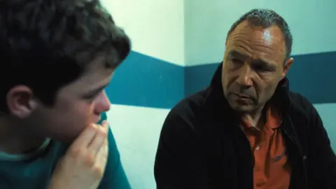 Netflix Owen Cooper, covering his mouth, while speaking to co-star Stephen Graham, in a scene from Adolescence