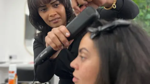 A woman with black hair has it straightened by another woman at a hair salon. The straightening irons are around strands of hair.