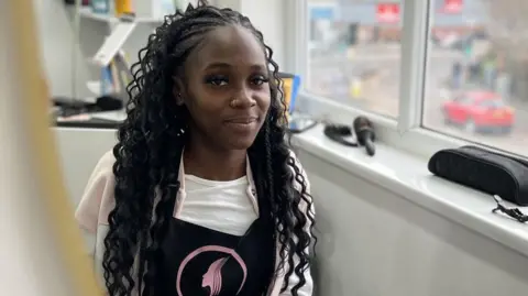 A woman with long black hair in waves looks at the camera with a slight smile. To her left is a window with a hairbrush and other equipment on the sill. She is wearing a white top under a pink jersey top and a black and pink apron.