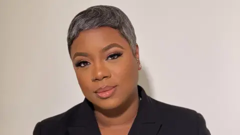 Erica Liburd A woman with short dark, grey hair looks into the camera with a slight smile. She wears a dark top with a black collar.