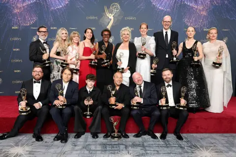 Getty Images The cast and crew of Adolescence pose with their Emmy trophies on a red carpet stage while wearing formal clothing.
