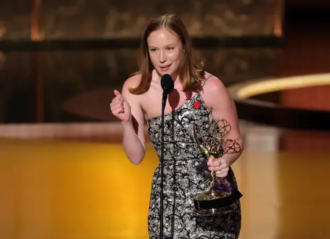 Getty Images Hannah Einbinder wears a sparkling gown with a red pin affixed to it as she speaks into a microphone onstage at the Emmy Awards. She holds her Emmy trophy for supporting actress in a comedy in her left hand.