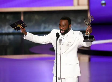 Getty Images Actor Trammell Tillman wears a white shirt and tuxedo and stretches his arms out while holding his Emmy Awards trophy in one hand and an envelope in another.