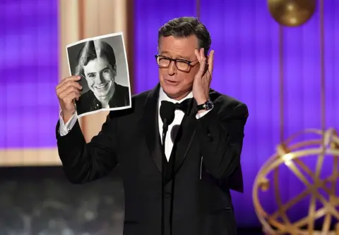 Getty Images Stephen Colbert wearing a tuxedo puts his hand on his cheek and holds up his old headshot onstage at the Primetime Emmy Awards