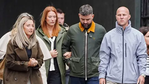 PA Media Jenni Stancombe, with long blonde hair and a brown coat, walks with her hands clasped together. David Stancombe, who has a bald head and is wearing a grey coat, walks a few paces from her.