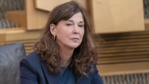 PA Media Dorothy Bain, who has long brown curly hair, sits in the Scottish Parliament chamber. She is wearing a blue suit jacket. 