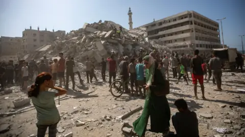 EPA Palestinians look for survivors in a large pile of rubble in the background. A girl covers her face with her hands in the foreground, while a woman walks past, looking down. 