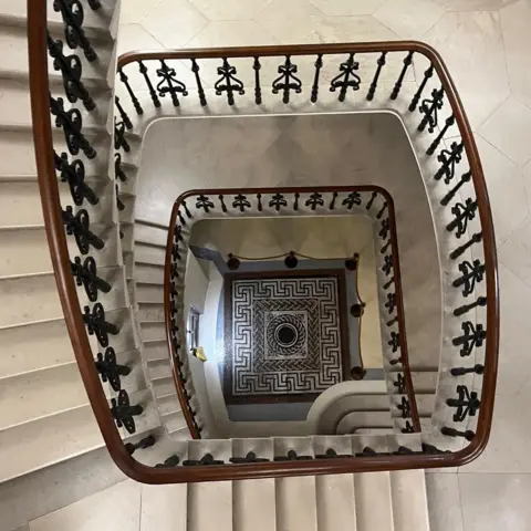A black, white and grey geometric mosaic at the bottom of a spiralling stone stair case which has a wooden bannister