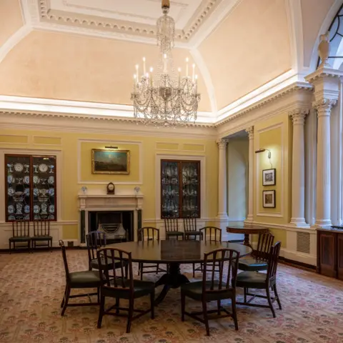 Bank of England Museum. Photo by Monica A Walker Vadillo A round wooden table is surrounded by wooden chairs in a yellow room which has white columns around it. A chandelier hangs above the table.