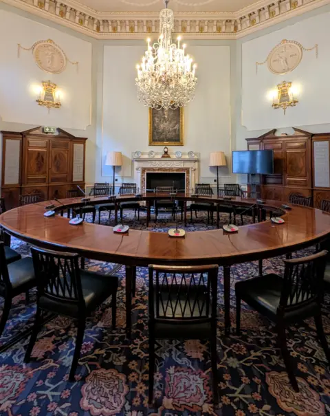 Bank of England Museum. Photo by Monica A Walker Vadillo Bank of England's Committee Room with wooden chairs surrounding a round table which has microphones placed around it. The room is octagonal with blue walls, a chandelier and decorated white and gold ceiling
