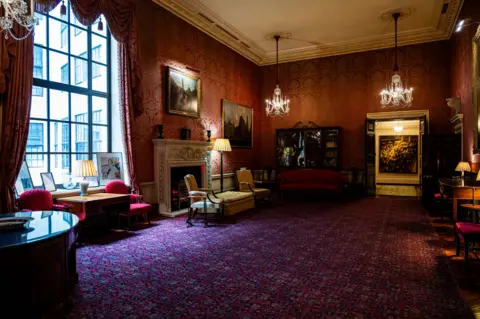 Bank of England Museum. Photo by Monica A Walker Vadillo Ante room in the Bank of England which has red and gold silk-covered walls, chandeliers hanging from the ceiling and a blue, red and purple patterned rug on the floor. Pictures hang on the wall and there is a decorative stone fireplace, with other furniture around the room